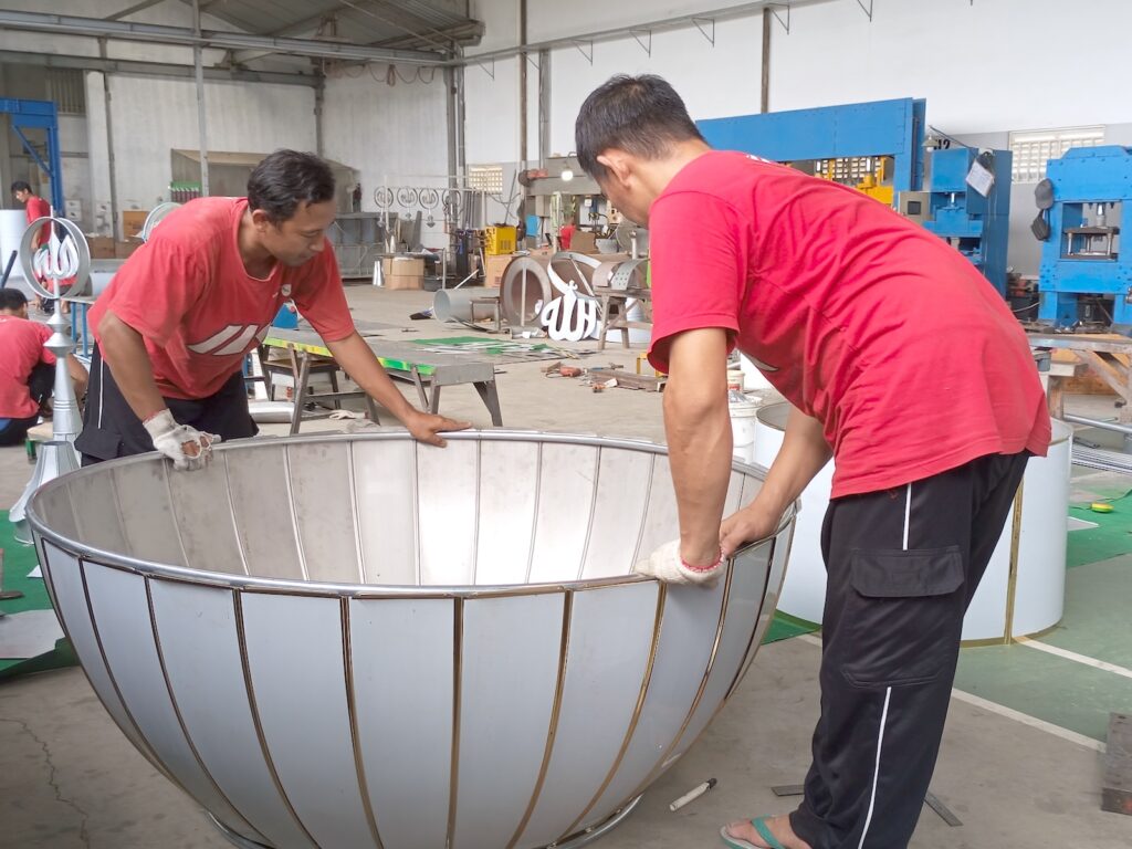 Tahapan Produksi Turbine Ventilator di Workshop PT Mustaka Multi Tehnik
Berdasarkan proses manufaktur yang terlihat pada workshop PT Mustaka Multi Tehnik, terdapat beberapa tahapan penting sebelum produk siap dipasarkan.
1. Persiapan Material Berkualitas
Produksi diawali dengan pemilihan material logam yang sesuai spesifikasi.
Material dipilih berdasarkan beberapa pertimbangan seperti:
ketahanan korosi,
kekuatan struktur,
kemudahan proses pembentukan,
serta umur pemakaian.
Pemilihan material yang tepat menjadi fondasi utama dalam menghasilkan turbine ventilator yang awet.
2. Pembentukan Komponen Menggunakan Mesin Produksi
Seluruh komponen diproses menggunakan berbagai mesin produksi untuk memperoleh dimensi yang presisi.
Pada tahap ini dilakukan:
pemotongan material,
pembentukan lengkungan,
proses press,
serta penyesuaian ukuran sesuai standar produksi.
Presisi dimensi sangat penting agar seluruh komponen dapat dirakit secara sempurna.
3. Proses Perakitan Manual oleh Teknisi Berpengalaman
Tahap berikutnya adalah proses assembling.
Pada gambar terlihat teknisi melakukan penyusunan panel-panel ventilator secara manual dengan penuh ketelitian.
Proses ini meliputi:
pemasangan panel luar,
penyatuan rangka,
pengecekan kesesuaian bentuk,
penyelarasan setiap sambungan.
Ketelitian pada tahap ini menentukan keseimbangan putaran turbine ventilator saat digunakan.
4. Pemeriksaan Kualitas (Quality Control)
Sebelum masuk tahap finishing, setiap unit melewati proses inspeksi kualitas.
Pemeriksaan dilakukan terhadap:
bentuk keseluruhan,
kekuatan sambungan,
kerapihan finishing,
kesesuaian ukuran,
serta fungsi mekanis.
Quality Control memastikan setiap produk memenuhi standar kualitas perusahaan sebelum dikirim kepada pelanggan.